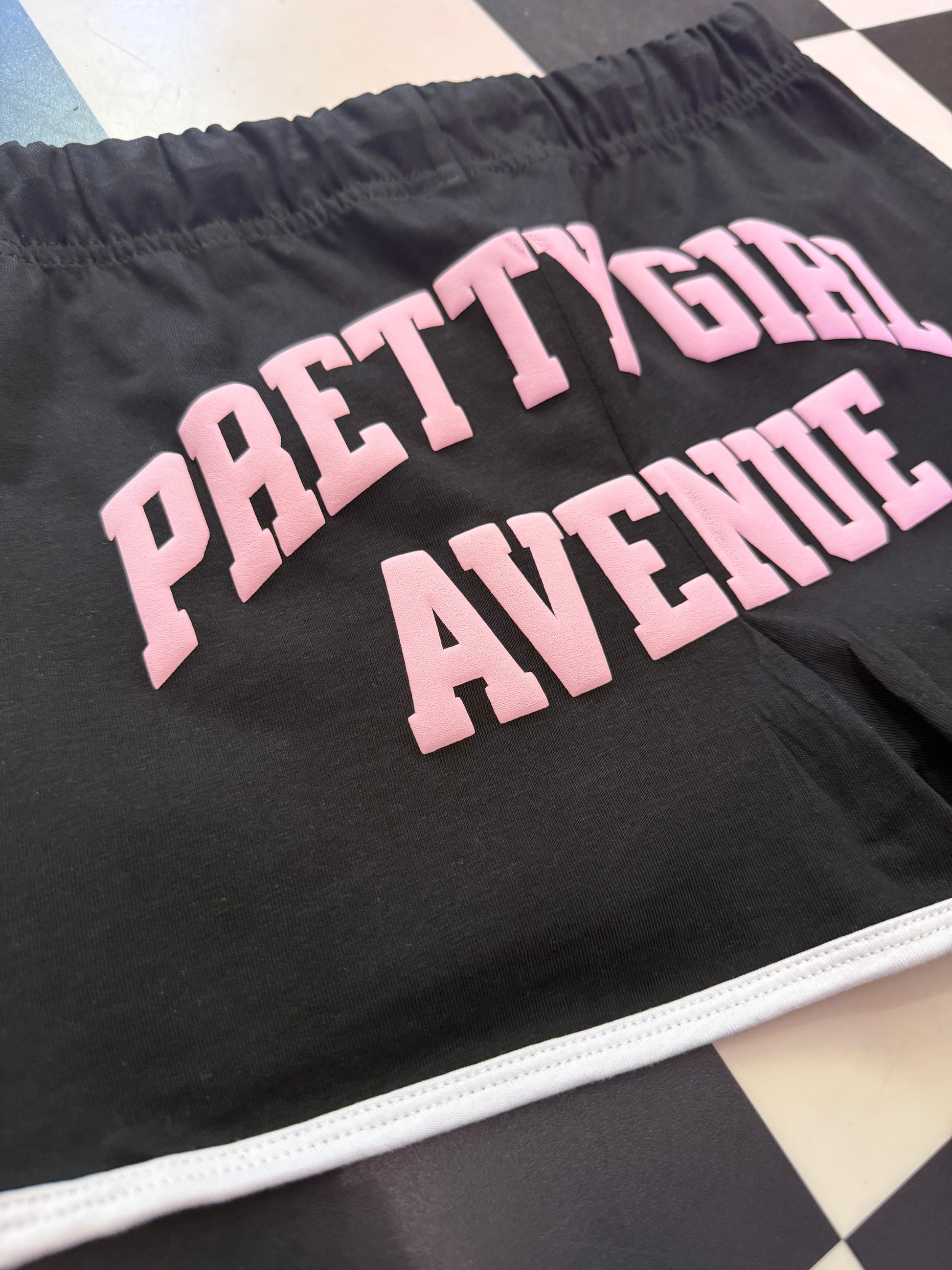 Black shorts with pink text 'PRETTY GIRL AVENUE' on a checkered floor.