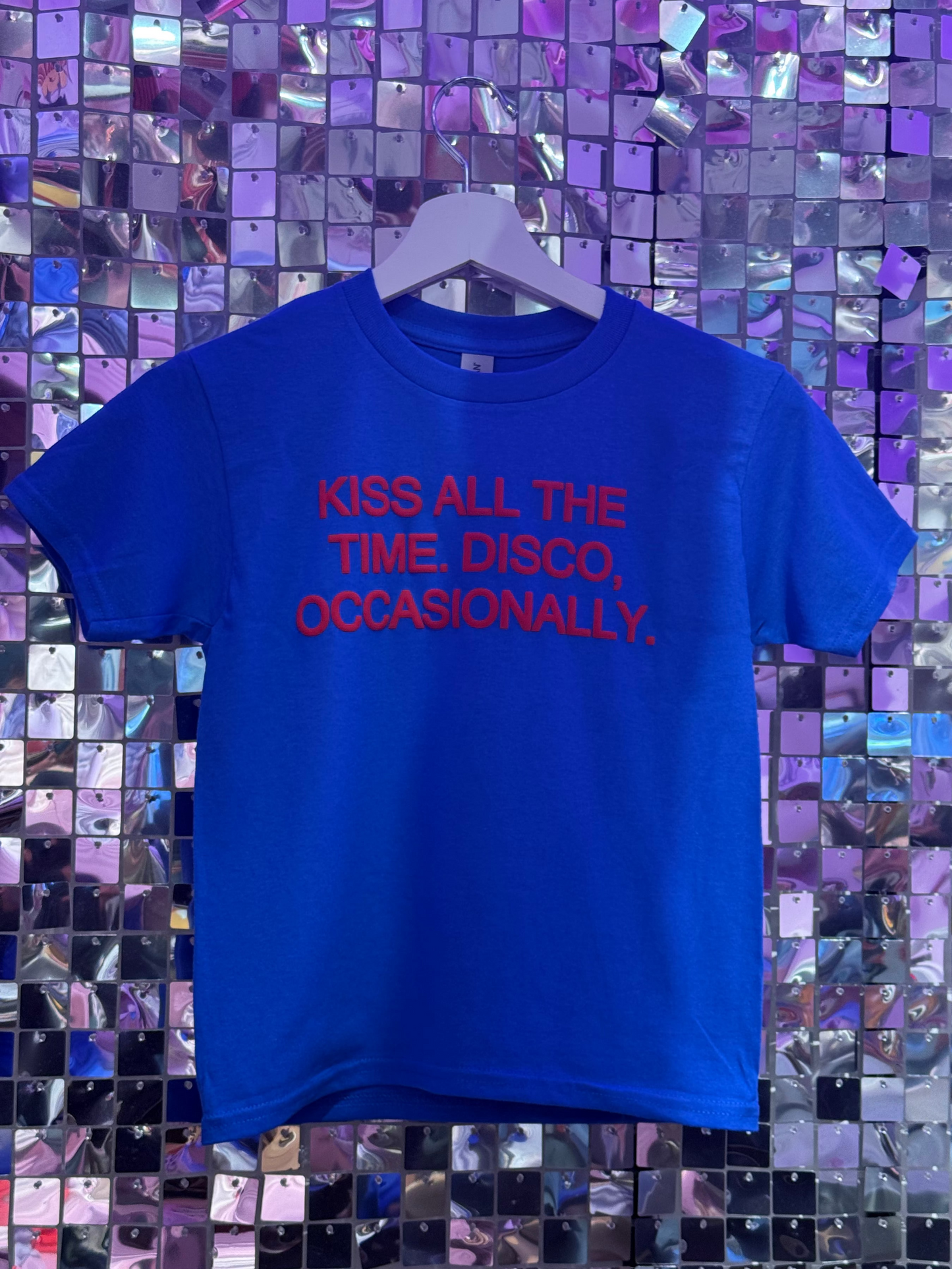 Blue t-shirt with red text 'KISS ALL THE TIME, DISCO, OCCASIONALLY.' on a hanger against a mosaic background.